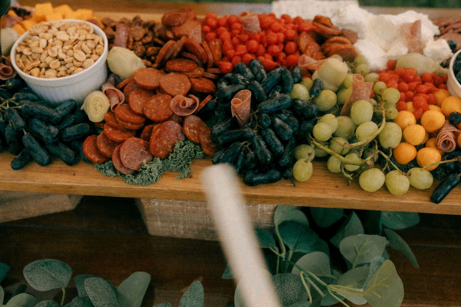 A wooden charcuterie board with meats, grapes, tomatoes, nuts, and dark vegetables at an outdoor event.