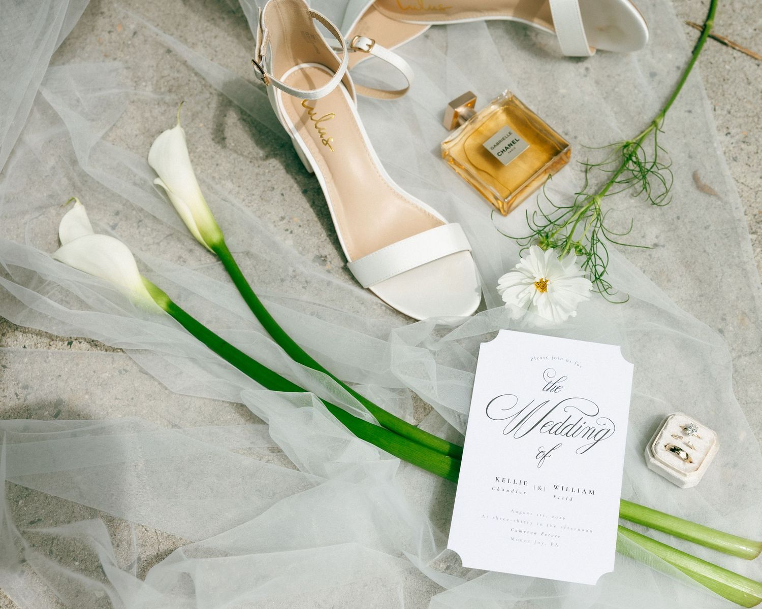 Elegant wedding flat lay with white heels, calla lilies, perfume, ring box, and wedding invitation on sheer veil.