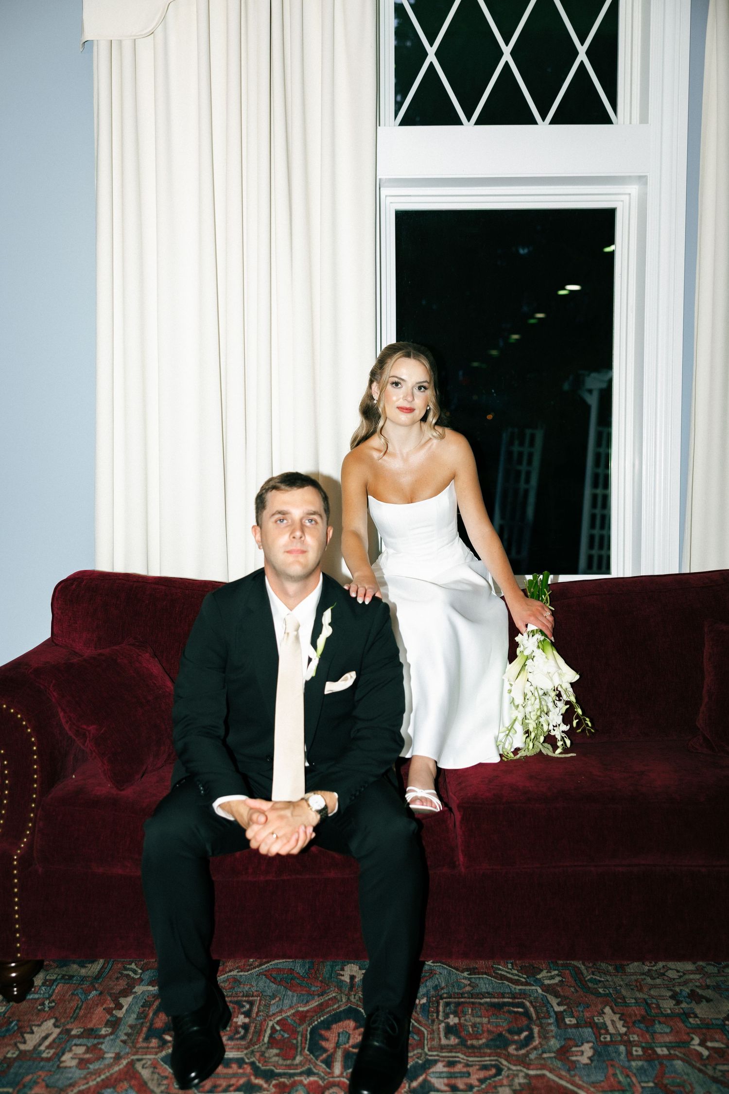 Bride in white gown holds bouquet beside groom in black suit seated on red velvet sofa near window.