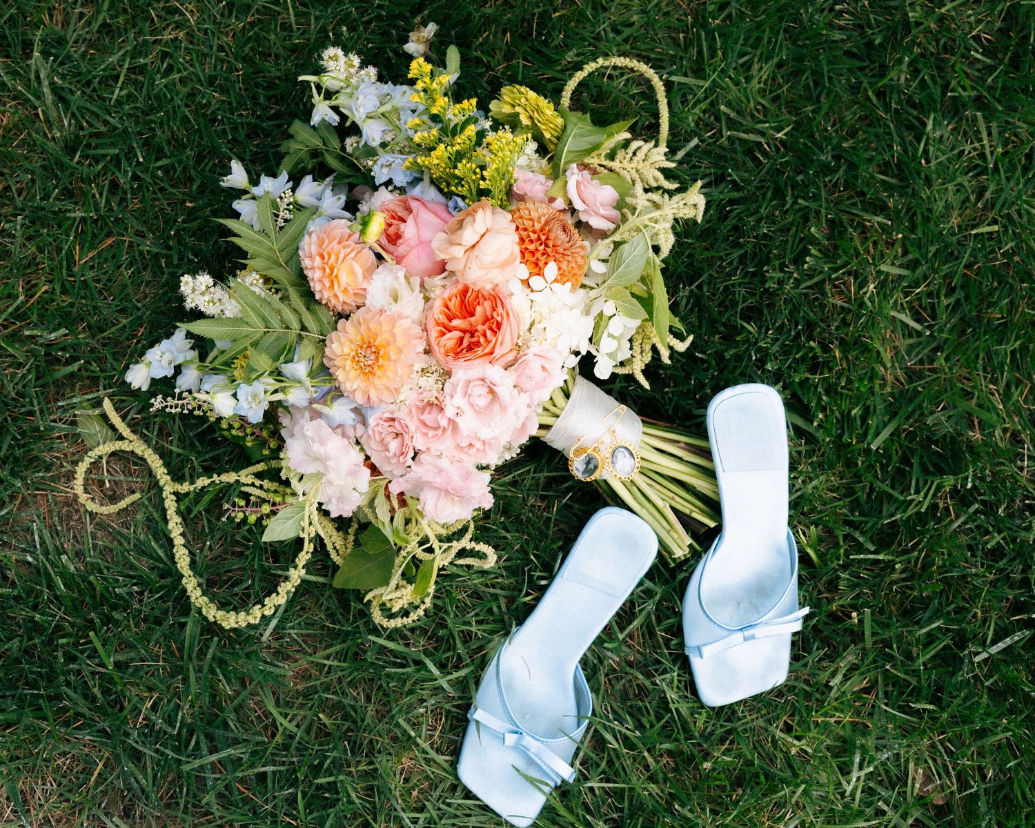 Bridal bouquet with peach and pink roses beside white satin heels on green grass.