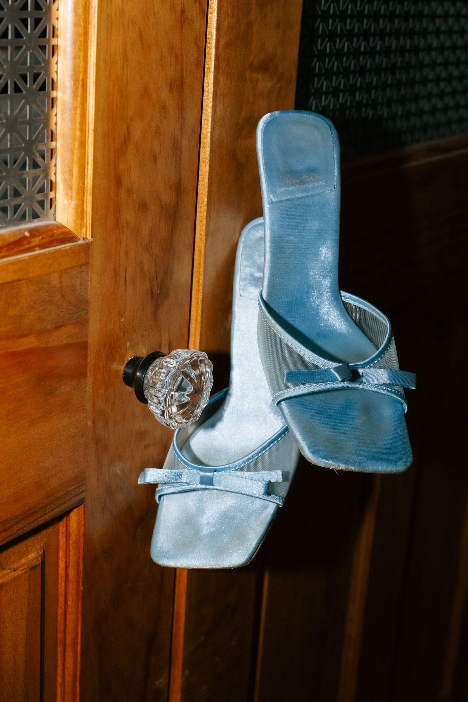 Light blue satin heeled mule sandals with bow detail hanging on a wooden door knob.