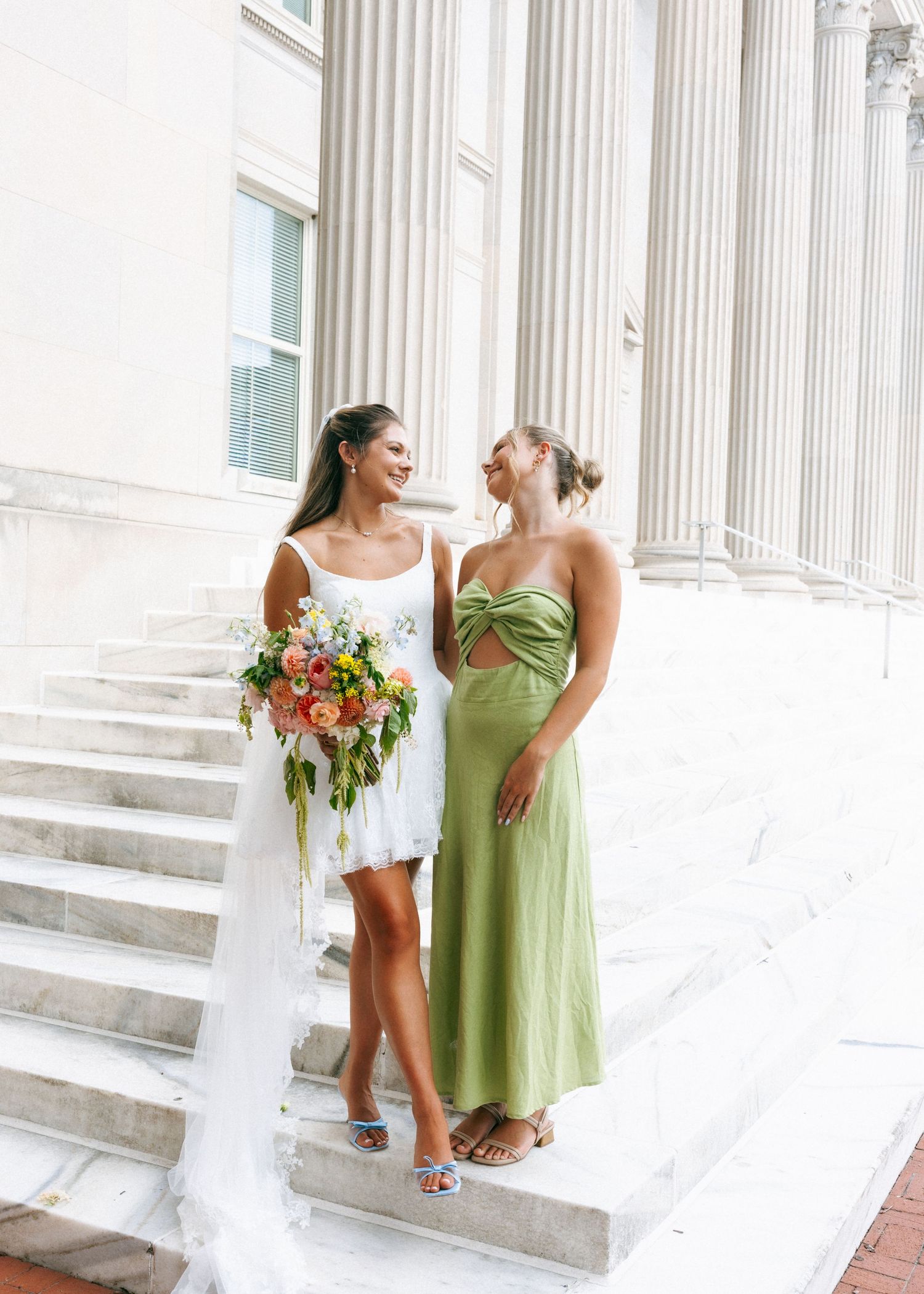 Bride in white mini dress holding colorful bouquet stands with bridesmaid in green satin dress on marble steps.