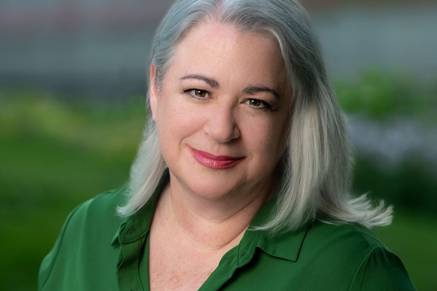 Professional headshot outdoors showing shoulder-length silver hair and emerald green blouse against soft-focus natural background.