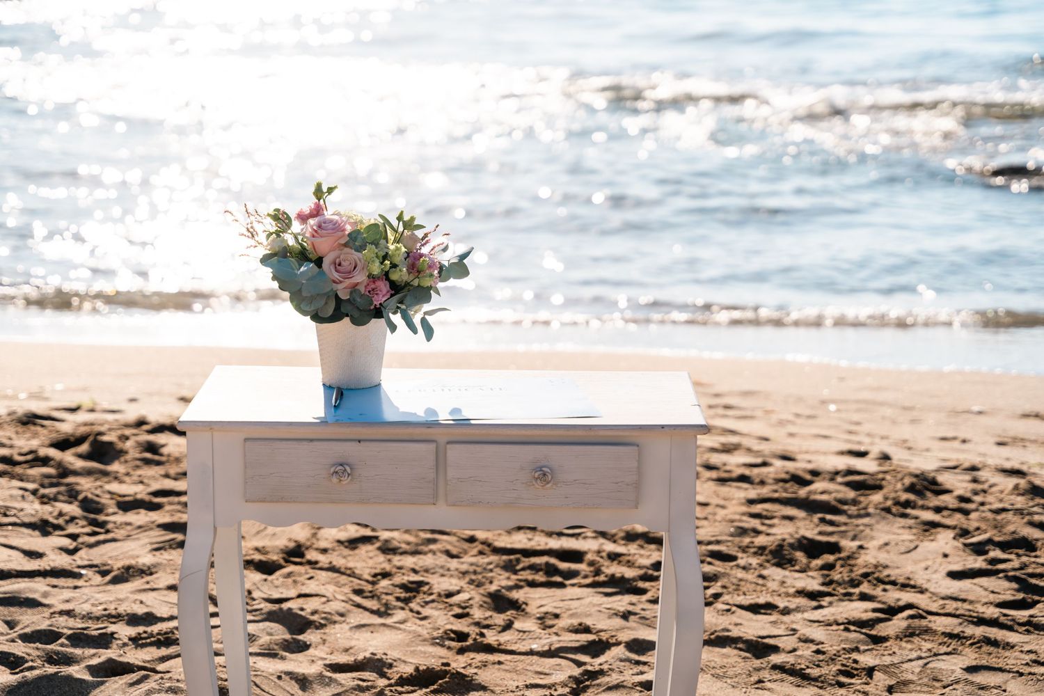 White vintage table with floral centerpiece arranged on sunny beach setting.