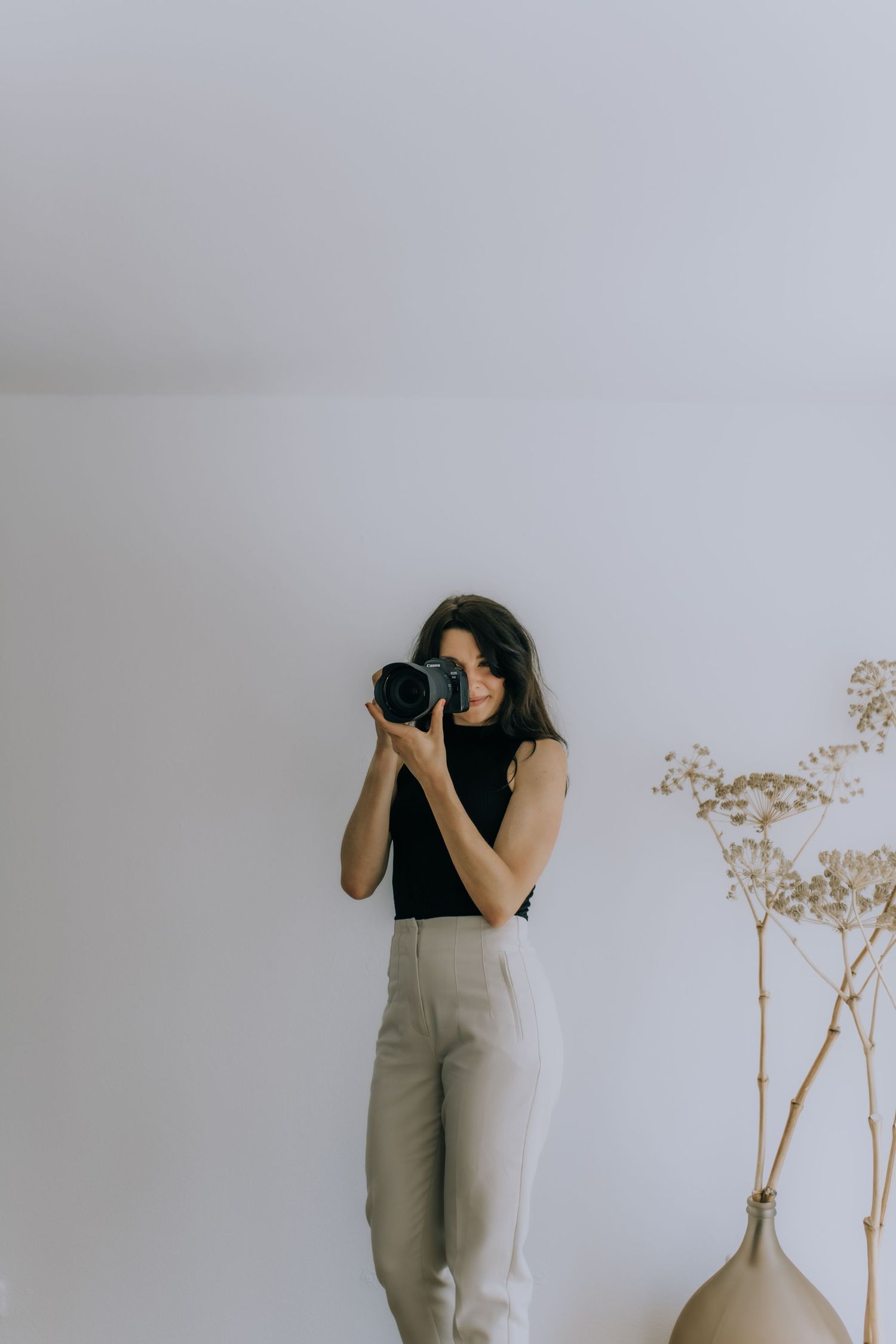 Photographer in black top and khaki pants captures images in minimalist white room setting.
