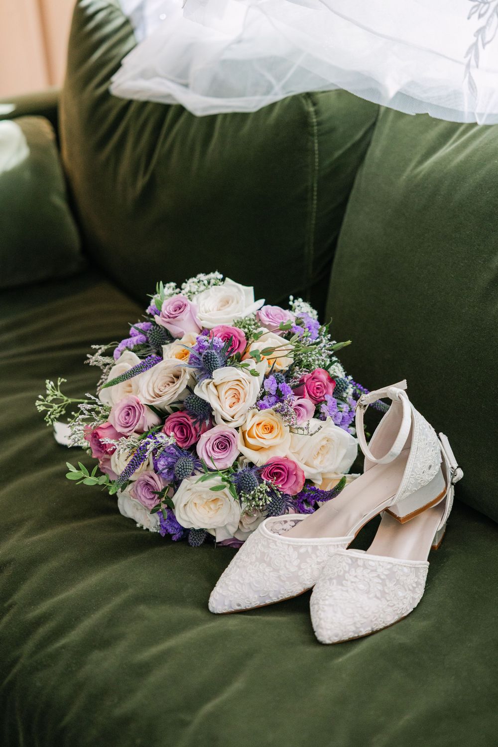 Elegant bridal bouquet with pink, purple, and cream roses beside white lace heels on a green velvet sofa.