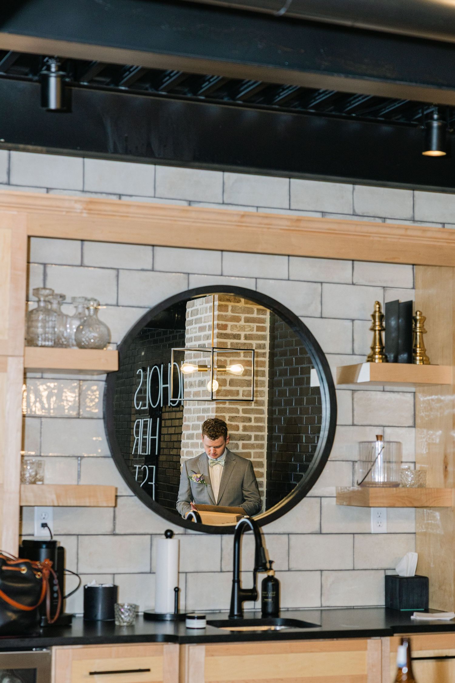 A man in a suit reflected in a round black-framed mirror above a kitchen sink with subway tile backsplash.