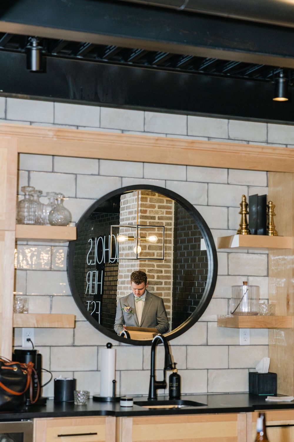A man in a suit reflected in a round black-framed mirror above a kitchen sink with subway tile backsplash.