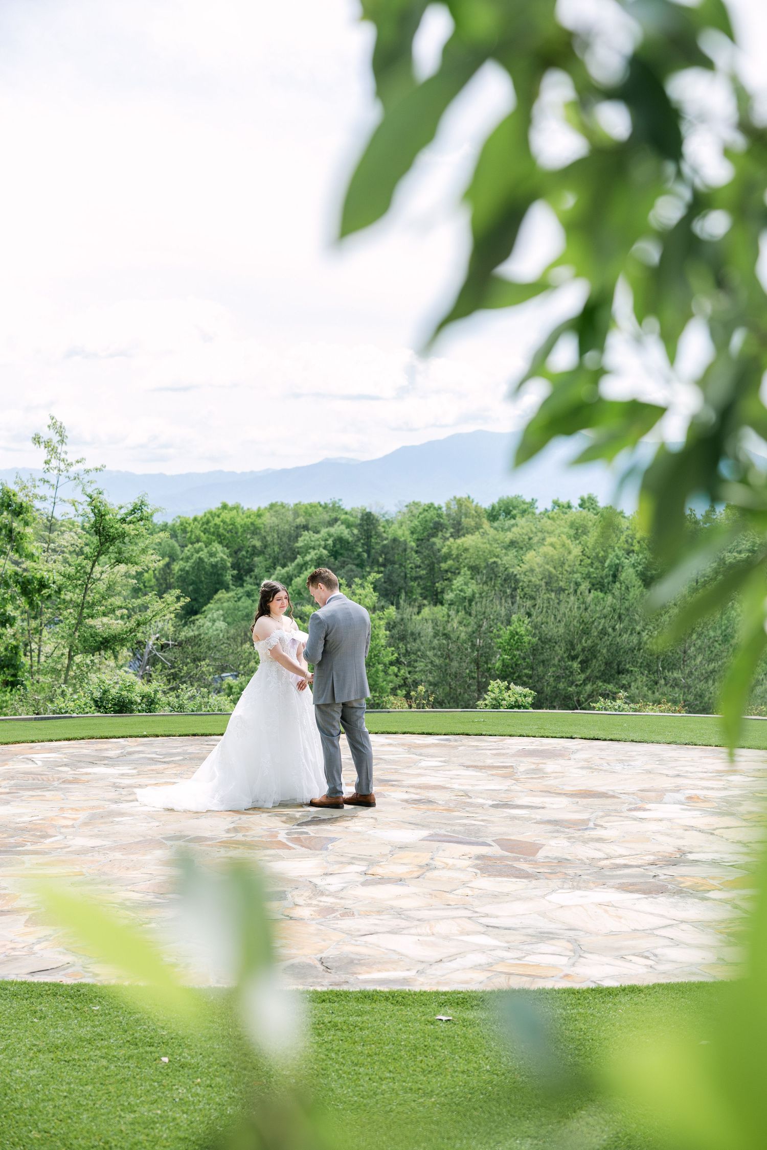 Bride and groom share a romantic moment on a stone patio surrounded by lush greenery and mountain views.