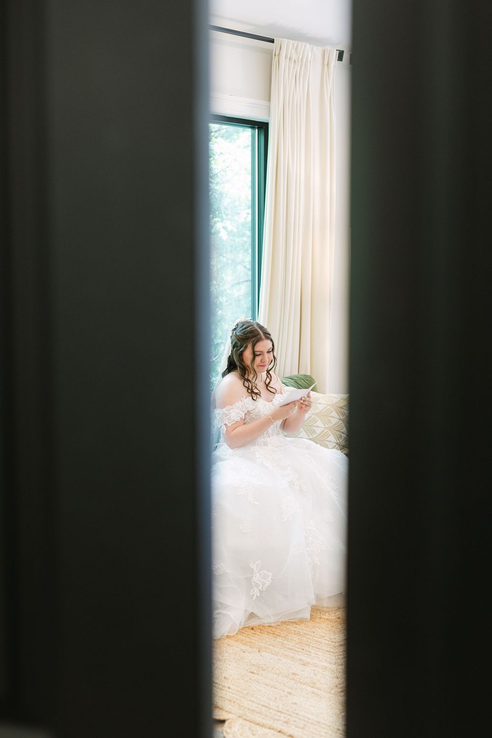 Bride in white gown reads a letter while seated by a bright window, captured through a doorway.