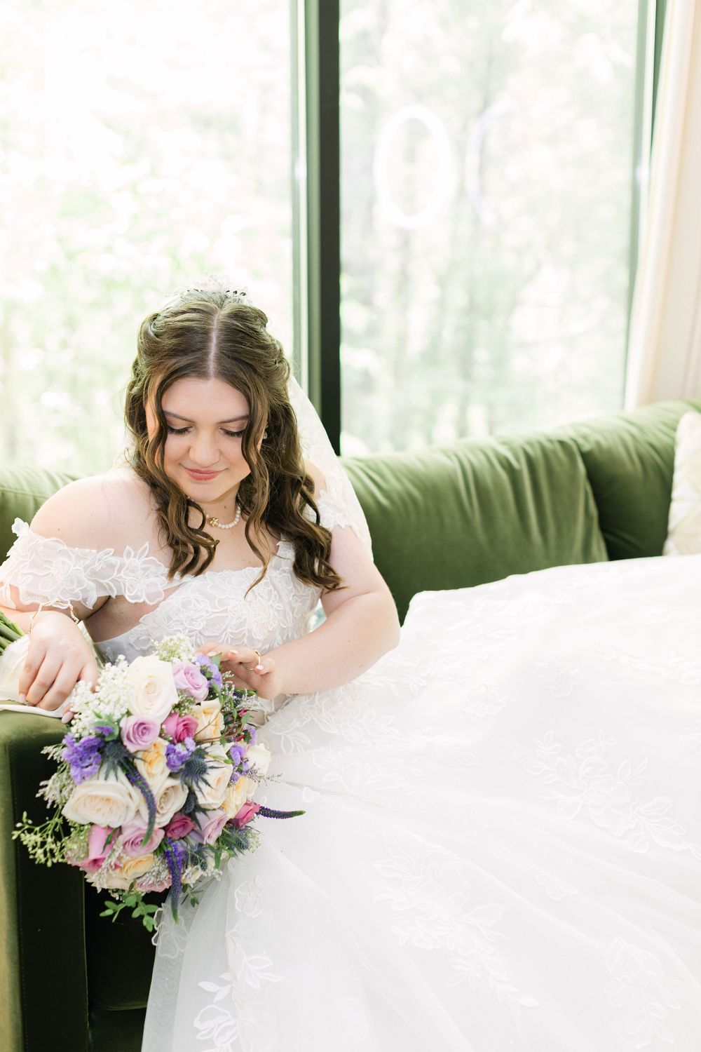 Bride in white lace off-shoulder gown sits on green velvet sofa holding colorful bouquet of purple and cream flowers.