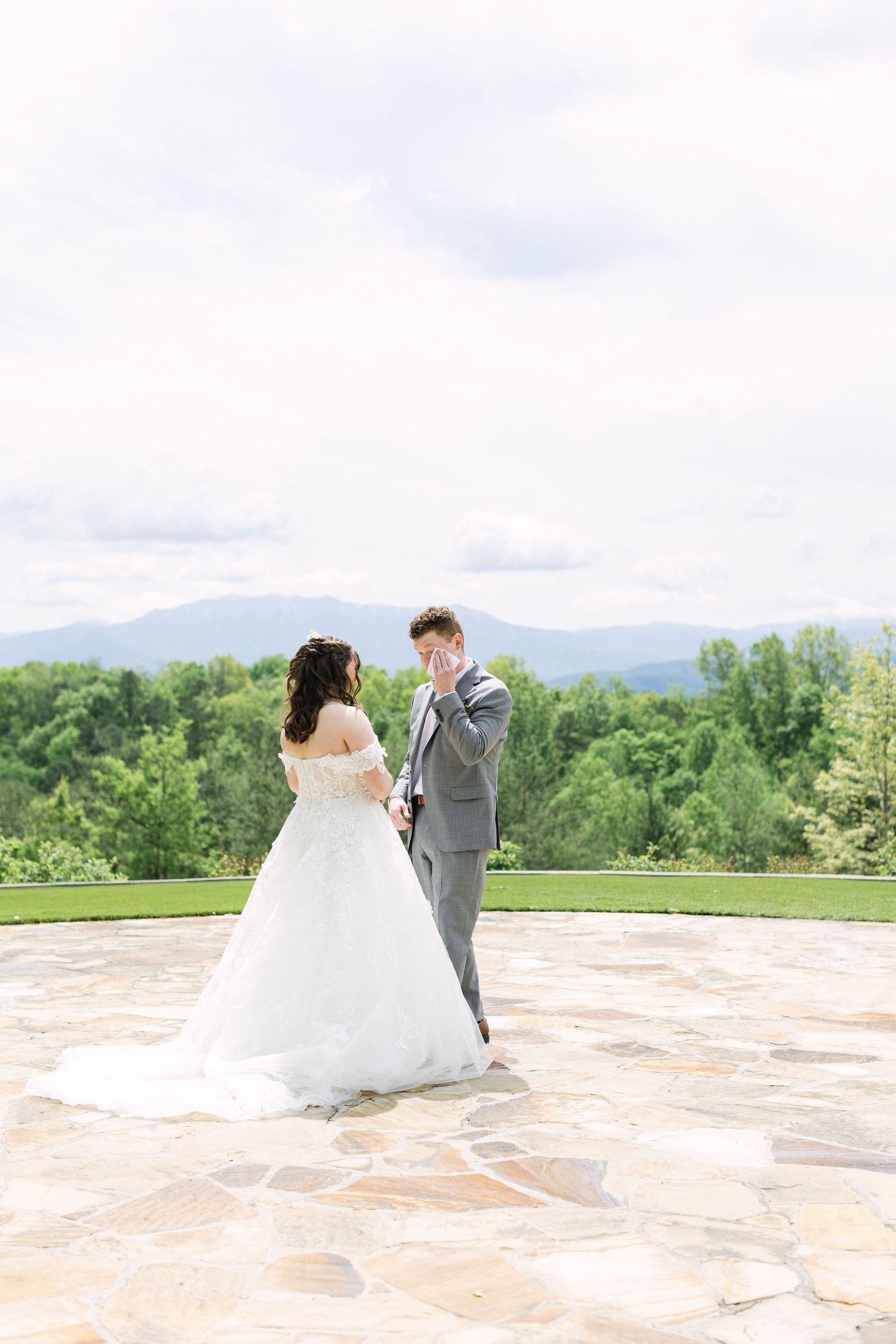 Groom wiping tears during first look with bride in white gown on stone patio with mountain views.
