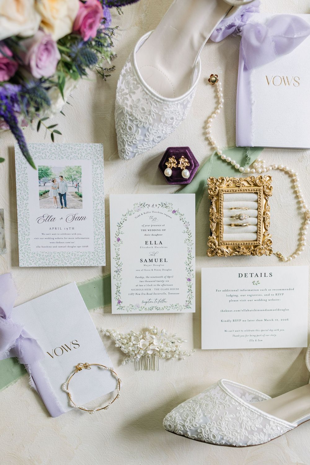 Elegant wedding flat lay featuring floral invitations, lace heels, pearl necklace, purple ring box, gold frame, and lavender vow books.