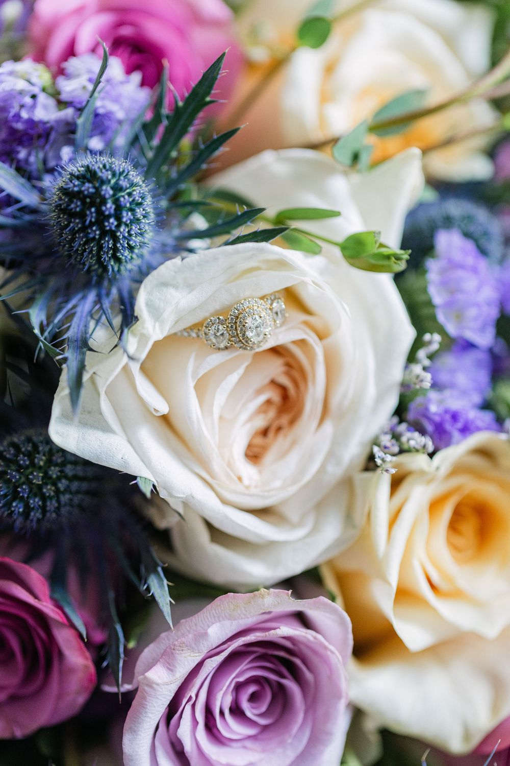 Diamond engagement ring nestled in a colorful bridal bouquet of cream, purple roses, and blue thistle flowers.