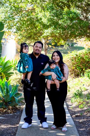 A family of four poses together on a garden pathway surrounded by lush greenery on a sunny day.