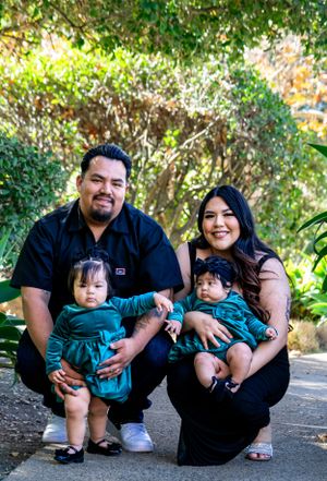 A family in coordinating teal and black outfits poses together on a garden path surrounded by lush greenery.