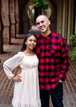 A father and daughter together outdoors wearing contrasting red plaid and white lace outfits against a brick wall background.