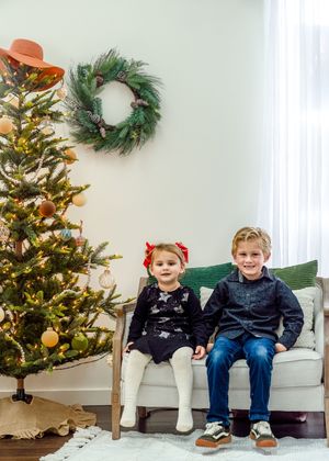 Two children sitting on green couch next to decorated Christmas tree and wreath.