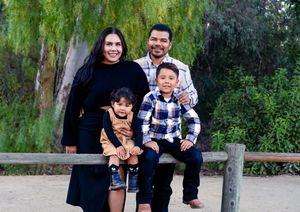 Family sitting together on rustic wooden bench surrounded by lush greenery in an outdoor setting.