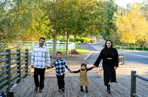 Family walking together on a paved path with autumn trees and white fencing in the background.