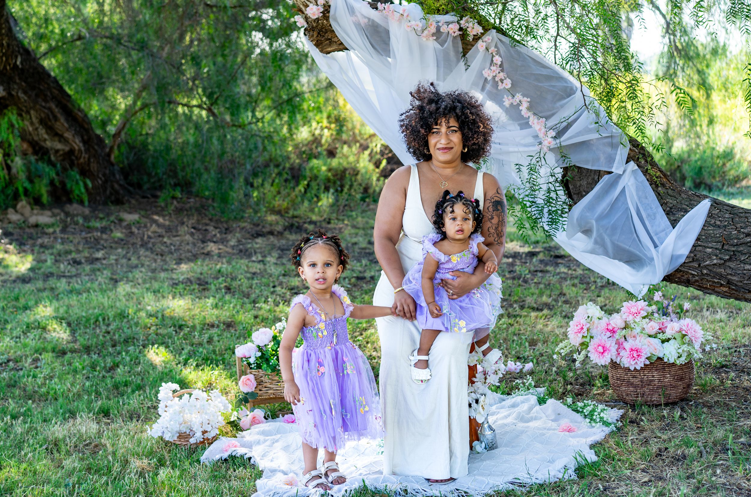 Woman in white dress poses outdoors with two young girls in purple dresses amid floral decorations and greenery.