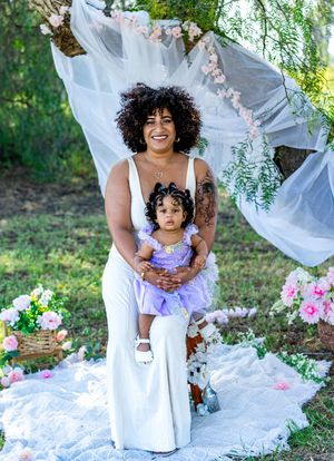 A woman in white holds a toddler in a purple tutu dress outdoors among pink flowers and flowing white fabric.