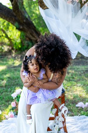 A mother with curly hair hugs her toddler daughter in a purple dress outdoors at a floral garden photo shoot.