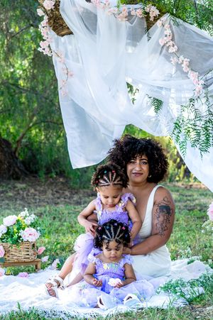 A mother in a white dress sits outdoors with two young girls in purple tutus under a floral canopy.