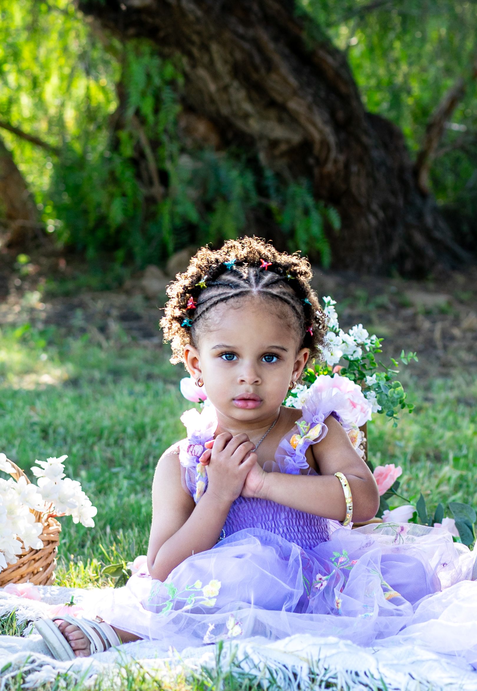 Young girl in purple tulle dress with floral crown sits on blanket outdoors surrounded by flowers and greenery.