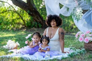 A mother in a white dress sits on a blanket outdoors with two young daughters in purple tutus amid flowers.