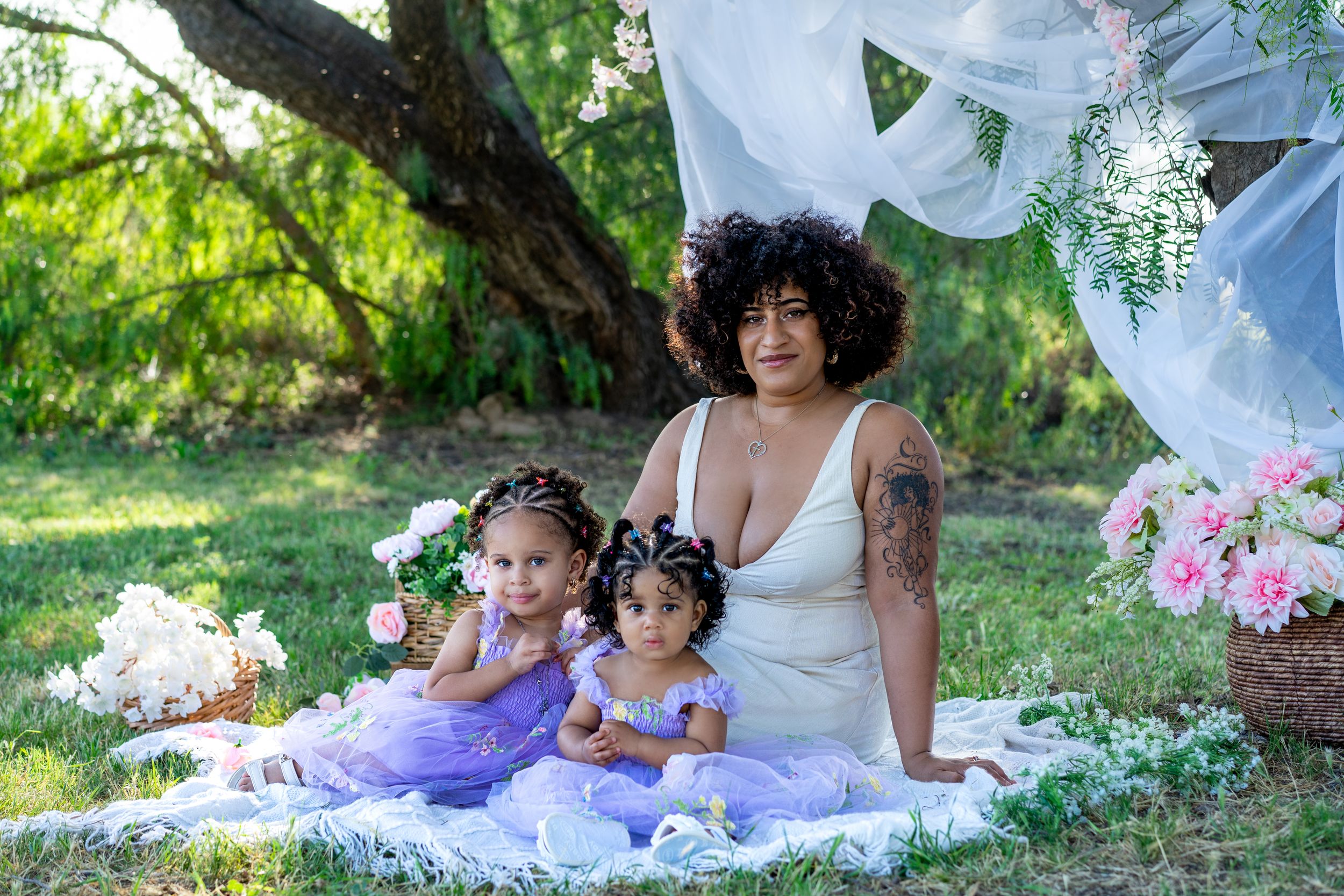 A mother in a white dress sits on a blanket outdoors with two young daughters in purple tutus amid flowers.