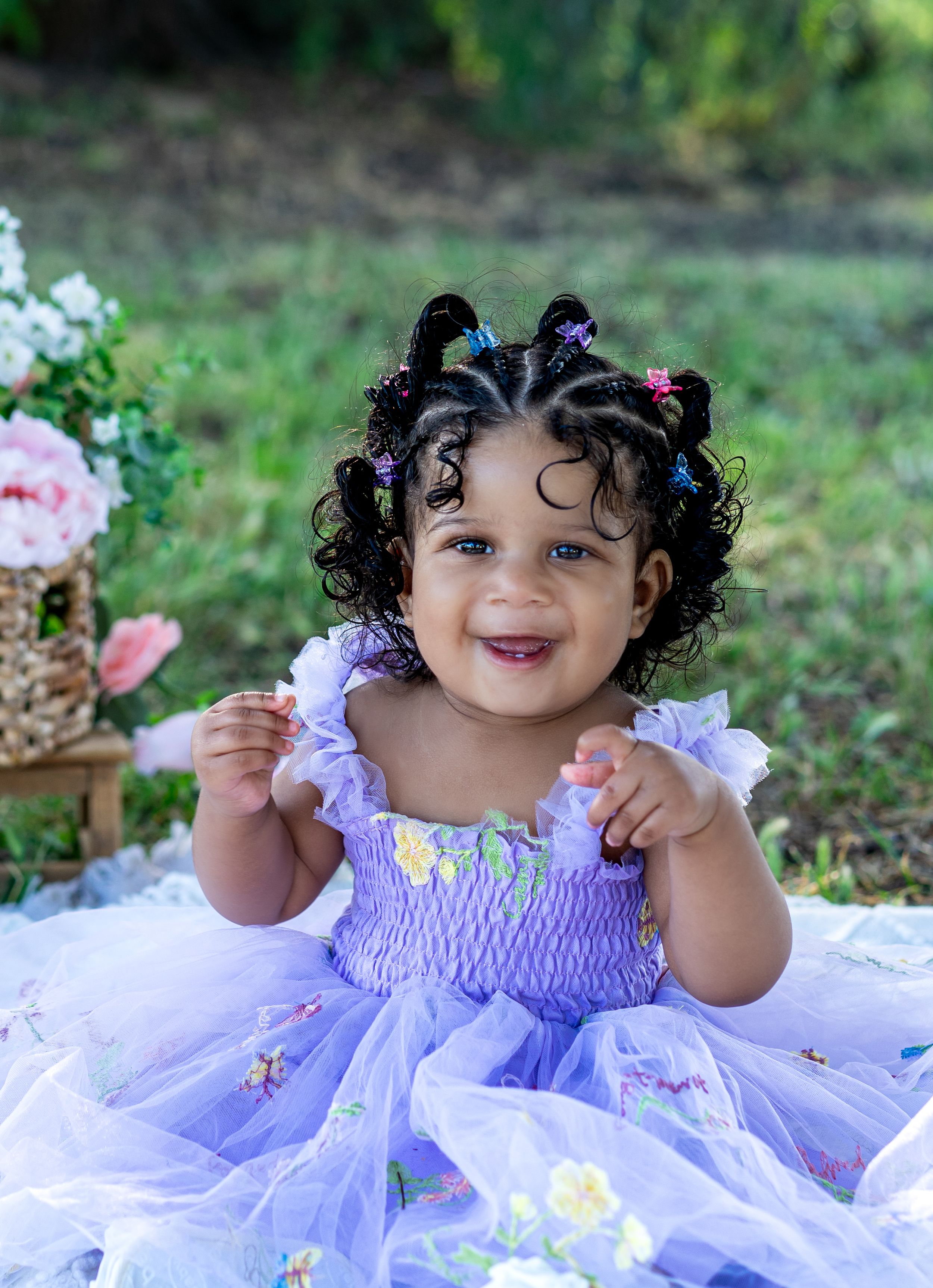 Smiling toddler girl in lavender tulle dress with curly hair and colorful clips sits outdoors among flowers.