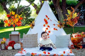 Autumn themed photo setup with white teepee tent, falling orange maple leaves, pumpkins and rustic decor in garden setting.