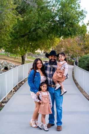 A happy family stands together on a walkway surrounded by green trees and railings.