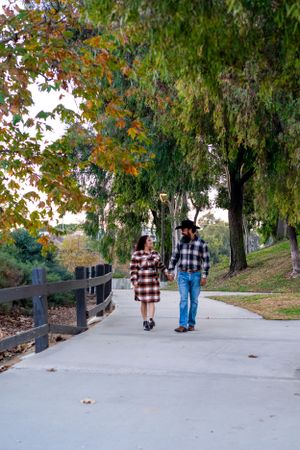 A couple wearing matching plaid shirts strolls down a tree-lined pathway on an autumn day.