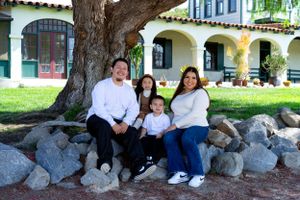 Family poses together on large rocks under a willow tree at a Spanish-style building.