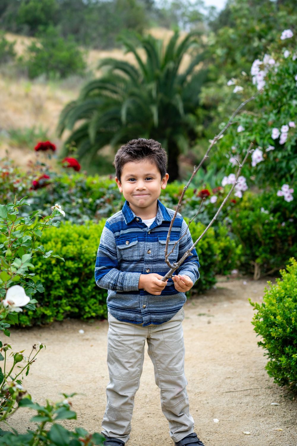 A young boy smiles while holding a fishing rod in a lush garden with colorful flowers and greenery.