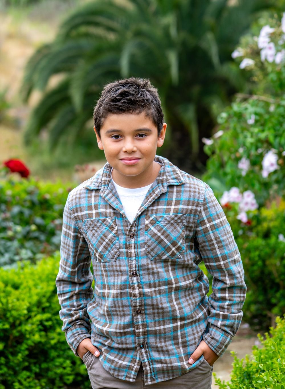 A young boy in a plaid shirt smiles confidently in a lush garden with green shrubs and colorful flowers.