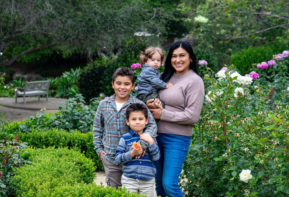 A mother poses with her three young children in a lush garden with white roses and green hedges.