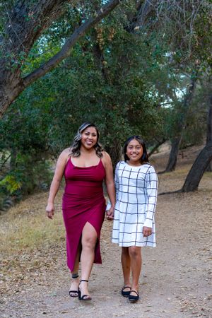 Two people walk together on a dirt path through trees wearing contrasting dresses - one burgundy and one white with grid pattern.