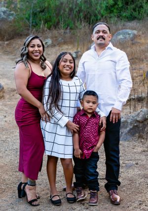 A family of four poses together outdoors in coordinated outfits including burgundy and white against a natural backdrop.