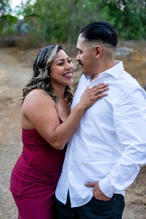 A romantic couple shares a joyful moment outdoors, with her wearing a burgundy dress and him in a white button-down shirt.