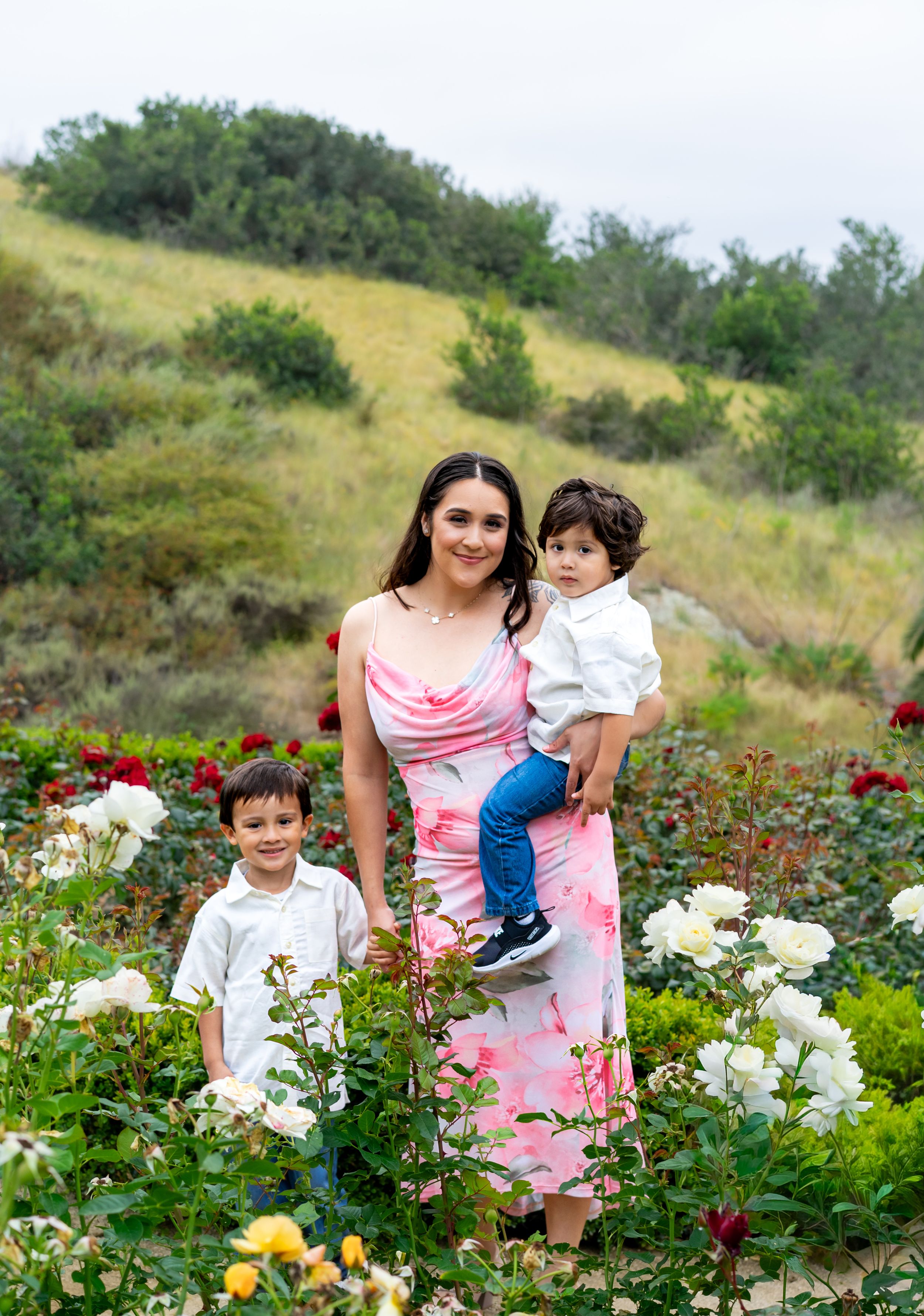 A woman in a pink floral dress holds a toddler while standing with a young boy in a colorful rose garden.