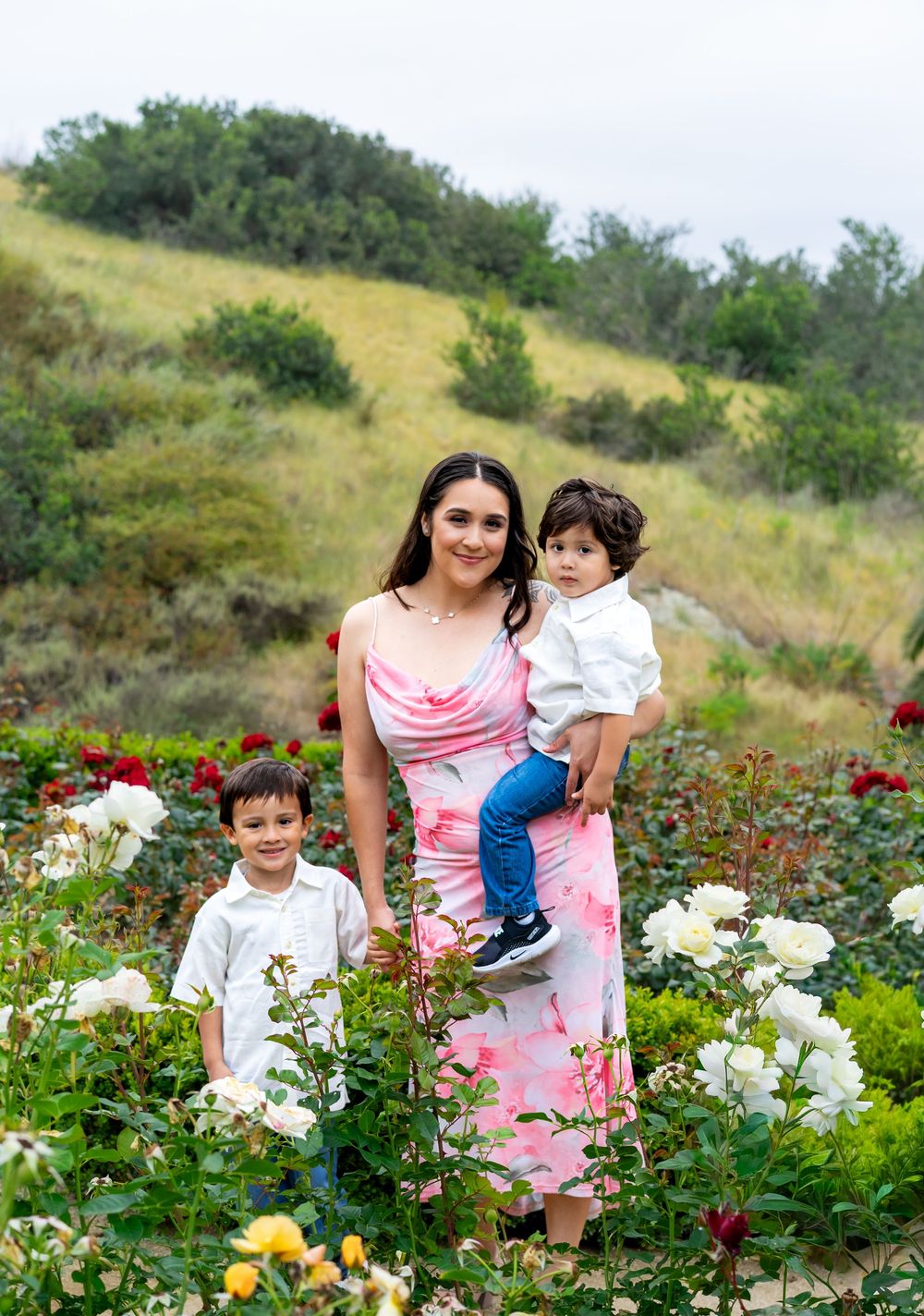 A woman in a pink floral dress holds a toddler while standing with a young boy in a colorful rose garden.