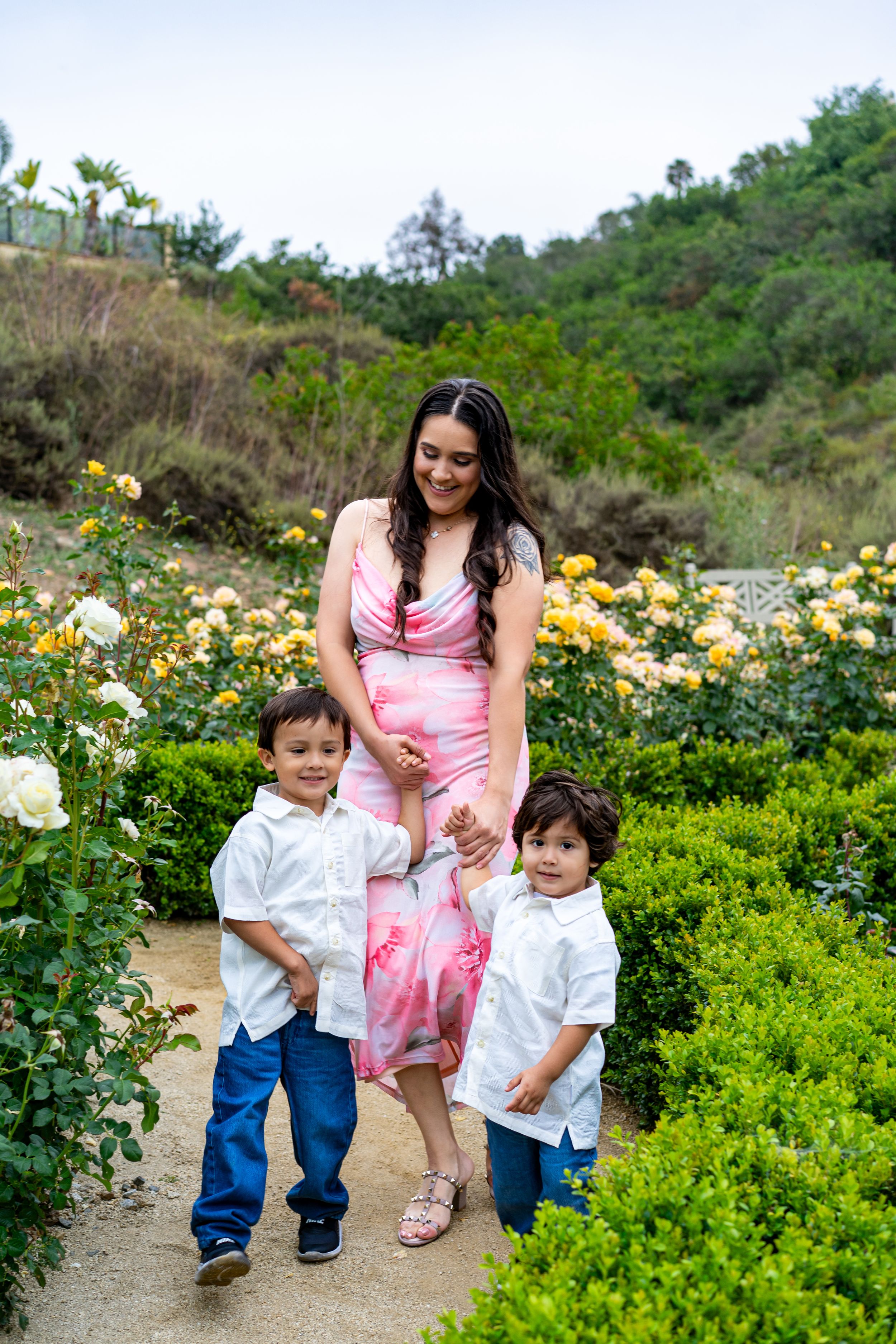 A woman in a pink floral dress poses with two young boys in white shirts and jeans in a rose garden.