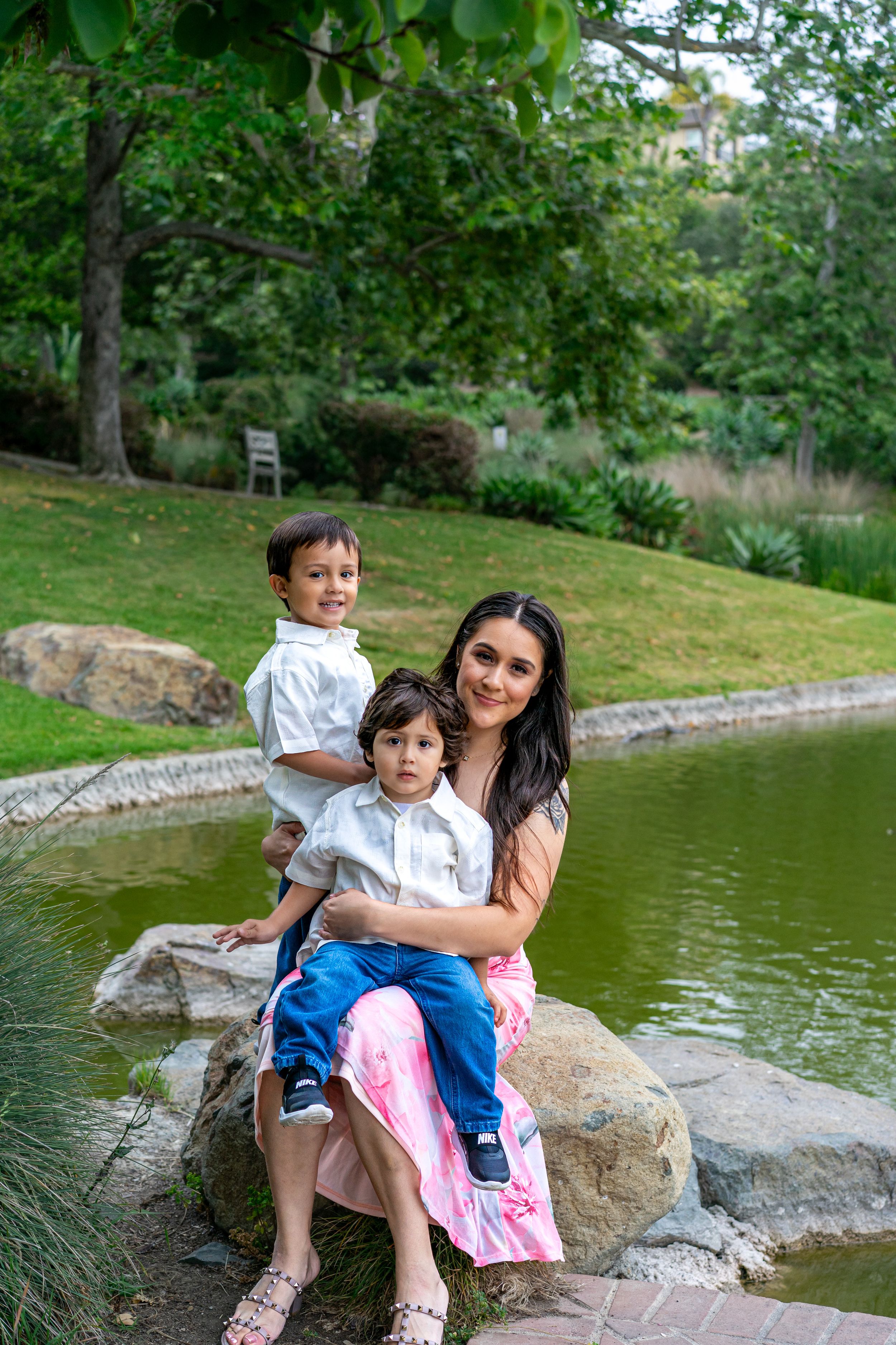 A woman in a pink dress sits on rocks by a pond, smiling with two young boys in a lush green park.