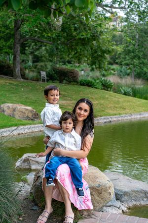 A woman in a pink dress sits on rocks by a pond, smiling with two young boys in a lush green park.