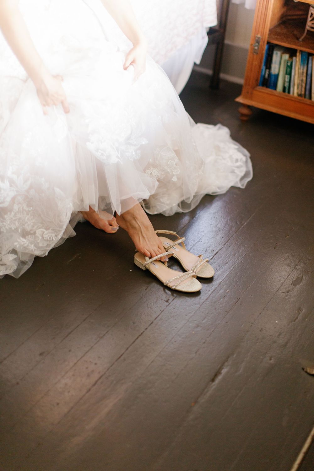 Bride in white lace wedding gown adjusting nude flat sandals on dark hardwood floor.