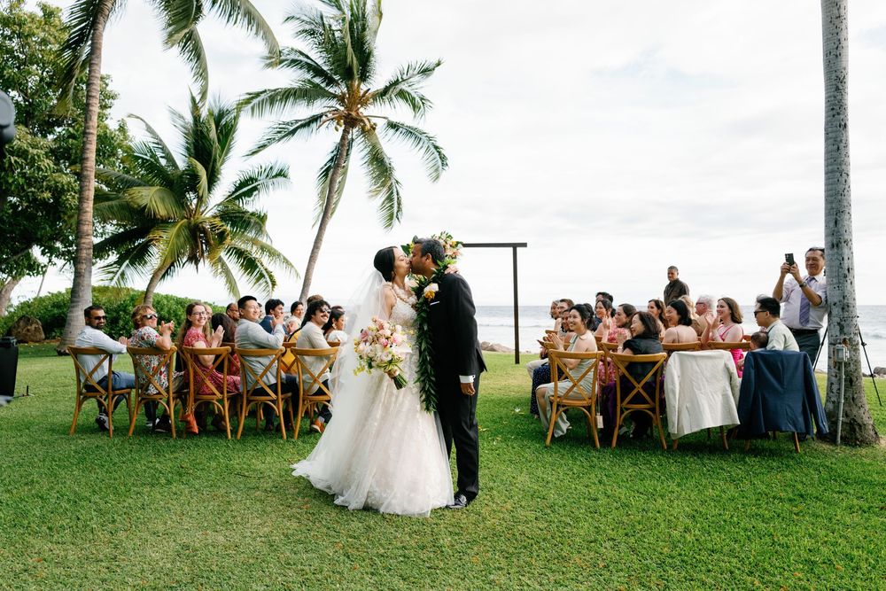 Couple shares first kiss at outdoor Hawaii wedding ceremony with guests seated at tables under palm trees at Olowalu Plantation House.