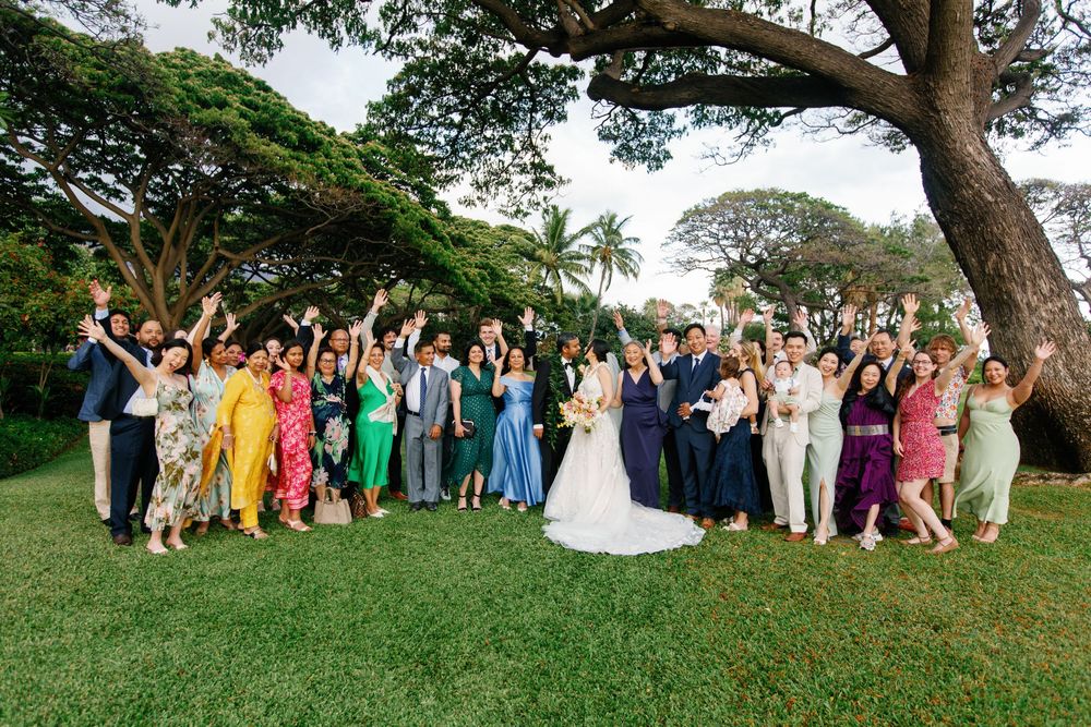 Large wedding party celebrates outdoors in Hawaii, guests in colorful attire cheering under lush green trees.